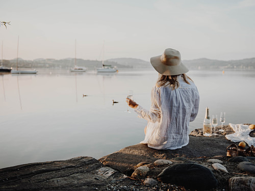 Woman with glass of sparkling wine sitting on rocky lake shore