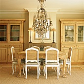 Freundliches Esszimmer mit hellen Holzvitrinen, geschnitztem Esstisch, weissen Vintage-Stühlen mit geflochtener Lehne und einem prunkvollen Kronleuchter