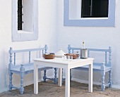 Sitzecke im Freien mit hellblauen Sitzbänken, weißem Tisch mit Tellerstapel, Tajine und Getränke