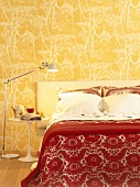 Ein Doppelbett mit roter Tagesdecke vor einer gelben Wand mit Pflanzenmotiv