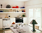 Simple, cheerful kitchen with crockery on long shelves and table lamp on white table