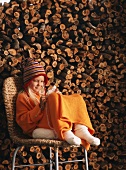 Girl sitting on chair knitting in front of stacked firewood