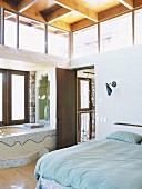 Doppelbett und mosaikbeflieste Badewanne in einem hohen Raum mit Holzbalkendecke und Oberlichtern
