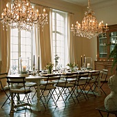 Gedeckter Landhaustisch mit Klappstühlen unter zwei prunkvollen Kronleuchtern in einem eleganten Saal mit raumhohen Fenstern