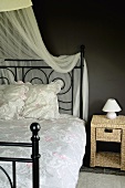 Klassisches Metallbett mit Betthimmel und romantischer Bettwäsche und ein Nachttischchen aus Rattan vor einer schwarzen Wand