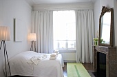 Schlafraum in Weiß mit Bett, Stehlampen, bodenlangen Fenstervorhängen & Kamin mit Spiegel