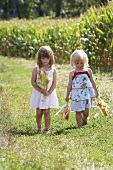 Two girls standing next to a corn field holding corn cobs