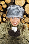 Young woman in winter hat holding cup, stack of wood behind