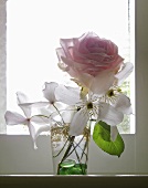 White flowers and pink rose in a glass by a window