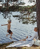 Junge springt in den See, Mutter lehnt sich am Baum an