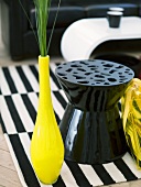 Gelbe Vase mit Ziergräsern neben schwarzen Plastikhocker auf schwarz weißem Teppich