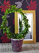 Wreath shaped plant in a pot and mirror in front of black wallpaper with a floral pattern