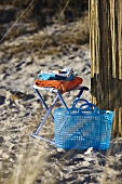 A blue plastic basket next to a stool with towel on a sandy beach