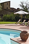 Clay planter beside a pool and lounge chairs under a sun umbrella with a view of a Mediterranean grange