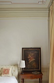 Corner of a bedroom -- a painting on a nightstand with a table lamp with a white shade