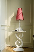 A table lamp with a red shade on a white occasional table with curved base standing against a wood panelled wall