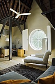 A white chaise longue in front of a round window with shutters in a converted attic with a dark wooden ceiling