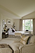 A light coloured, upholstered armchair with a footstool in a spacious bedroom with a double bed in front of a window
