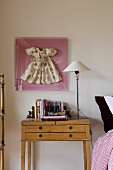 A table lamp with a white shade on a light wood bedside table with a framed doll's dress hanging on the wall