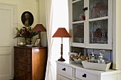 A white crockery cupboard and a table lamp with a red shade in the corner of a dining room