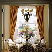A set table in front of a window with pleated, yellow curtains with a chandelier hanging in the middle