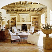 A view through a rounded archway in a Mediterranean living room with a rustic wood beam ceiling and white armchairs in front of a natural stone fireplace
