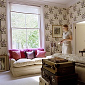 A stack of trunks, a light upholstered sofa with cushions in front of a window, patterned wallpaper and a woman in the room