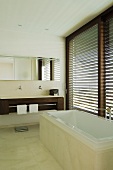 A designer bathroom - a bathtub in front of a window with closed blinds and a wooden washstand with a mirrored cabinet