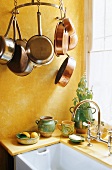 A yellow painted kitchen corner with hanging copper pans and a sink in front of a window