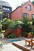 A red-painted house with a flight of stairs and a mini pool in a green courtyard with a wooden terrace