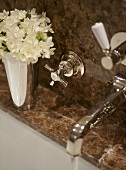 A detail of a traditional bathroom, showing a fitted bath with marble surround, chrome taps, water running, flowers in silver vase