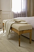 A knitted shawl on an antique bench with a light-coloured upholstered seat in a room with white-painted floorboards