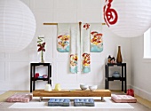 Japanese sitting room with a white panelled wall, hanging kimono, paper lanterns, black lacquered side tables, and a low wooden table with floor cushions.