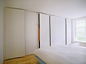 A modern, white minimalist bedroom with double bed, fitted wardrobes, wooden floor, plain window,