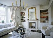 A traditional, neutral sitting room, with fireplace, gilt mirror, book shelves and modern glass coffee table
