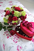 An open box of chocolates and a bunch of flowers on a floral patterned tablecloth