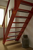 Red wooden stairs in front of a wooden wall and a milk can under the stairs