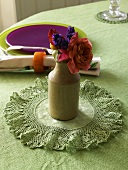 A mini bunch of flowers in a ceramic vase on crocheted mat with a coloured place setting