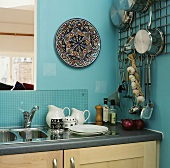 Küchenecke mit türkisen Wänden mit Keramikteller an einer Wand, an einem Metallgitter hängenden Küchenutensilien und einer Edelstahl-Spüle