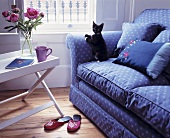 Schwarze Katze auf blauer Couch mit Muster neben weißem Beistelltisch