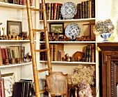 Bambusleiter und antiker Stuhl vor Bücherregalen mit Holznippes und alten Büchern in traditionellem Wohnzimmer