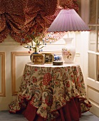 Brennende Lampe mit fliederfarbenem Plisseeschirm auf rundem Tisch mit floraler Decke