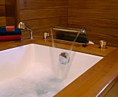Fliessendes Wasser aus flachem Wasserhahn und Duschkopf in hölzernem Wannenrand in Badezimmer mit Holzvertäfelung