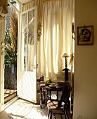 Sonne durch halboffene Terrassentür auf kleinen Sitzplatz mit antikem, französischem Holzmöbel vor weißem Vorhang