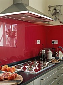 Spritzschutz aus rotem Glas über schwarzer Arbeitsfläche mit Gaskochfeld und Dunstabzugshaube aus Edelstahl
