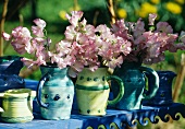 Rustikale Keramikkrüge in Blautönen mit pastellvioletten Wickensträussen auf blauem Gartentisch mit geschnitztem Zierrand