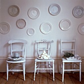 Weisses Arrangement von an der Wand aufgehängten Porzellantellern über antiken Holzstühlen mit Küchendekorationen