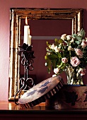 Spiegel in abgeschabtem, goldenem Bilderrahmen hinter Metall-Kerzenständer mit brennender Kerze und Rosenstrauss