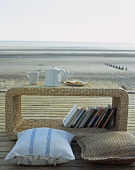 Ein Korbtisch mit Kissen und Büchern auf einem Holzdeck am Strand