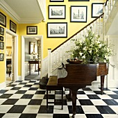 Brauner Flügel in Eingangshalle vor weisser Holztreppe mit Bildersammlung an hellgelben Wänden und Schachbrett Boden